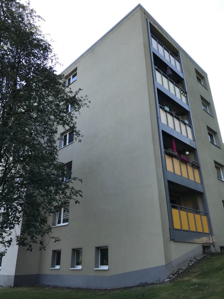 Ein fünfstöckiges Wohnhaus mit beigen Wänden und gelben Balkonpaneelen sowie einer sauberen Fassade steht neben einem belaubten Baum und einer Rasenfläche. Das Foto ist von der Seite aufgenommen, wodurch die Höhe des Gebäudes hervorgehoben wird.