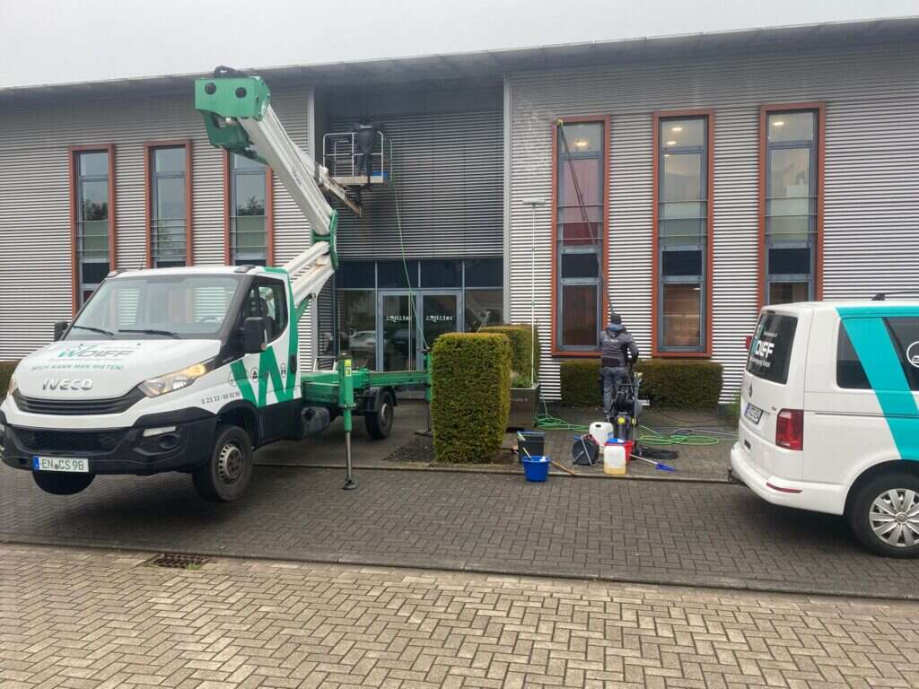 Zwei Mitarbeiter reinigen mit Hubsteiger und Hochdruckreiniger die Fassade und Fenster eines grauen Gebäudes