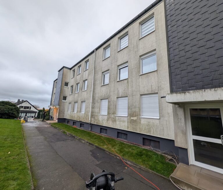 Mehrstöckiges graues Mehrfamilienhaus mit Rollläden und Gehweg